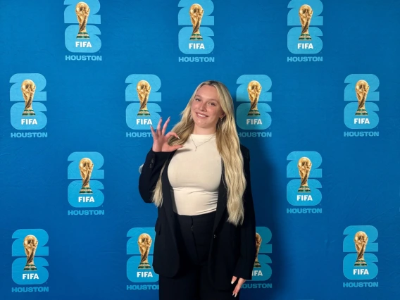 Samantha standing in front of a FIFA back drop. She's displaying the WC ("wildcats") had signal