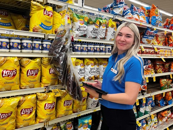 woman in blue shirt and dark pants standing in front of chip aisle in grocery store