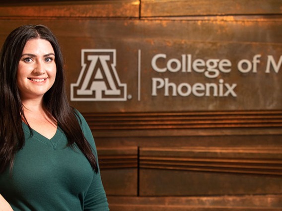 Chelsea Meraz stands smiling in a green shirt in front of a brown wall with the words "College of Medicine Phoenix."