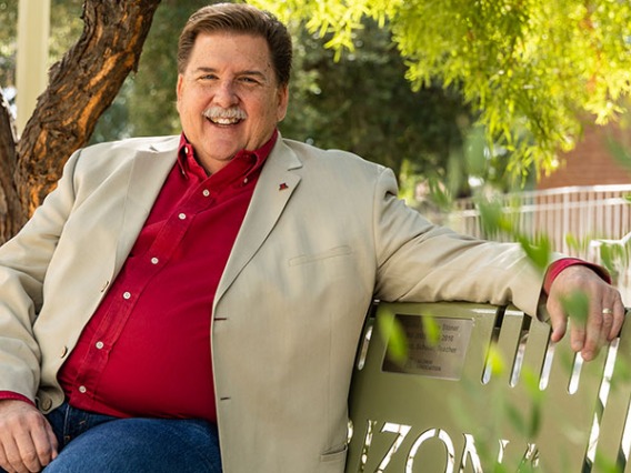 Jim Wells, in a red shirt and tan blazer, smiles while sitting on a green bench.