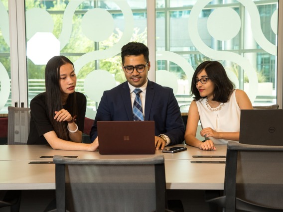 Three students sitting at table looking and pointing at laptop