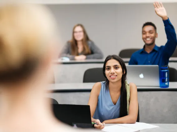 Three students in classroom facing teacher. One student has their hand raised.