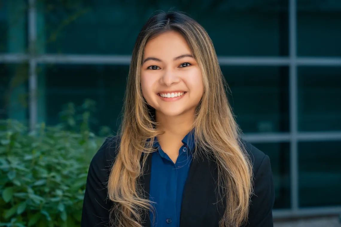 woman with long hair, blue shirt and dark blazer smiling in front of windows