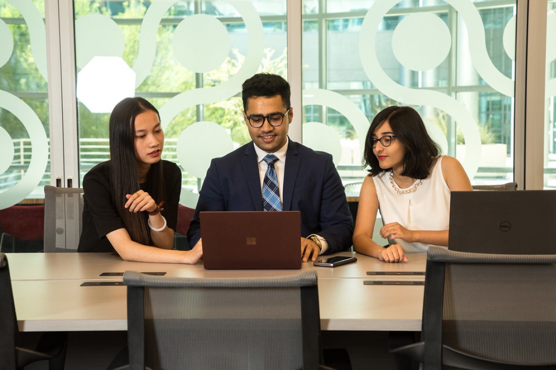 Three students sitting at table looking and pointing at laptop