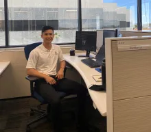 man with dark hair in white shirt looking at the camera while sitting at a desk