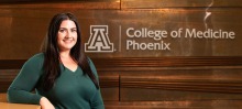 Chelsea Meraz stands smiling in a green shirt in front of a brown wall with the words "College of Medicine Phoenix."