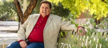 Jim Wells, in a red shirt and tan blazer, smiles while sitting on a green bench.